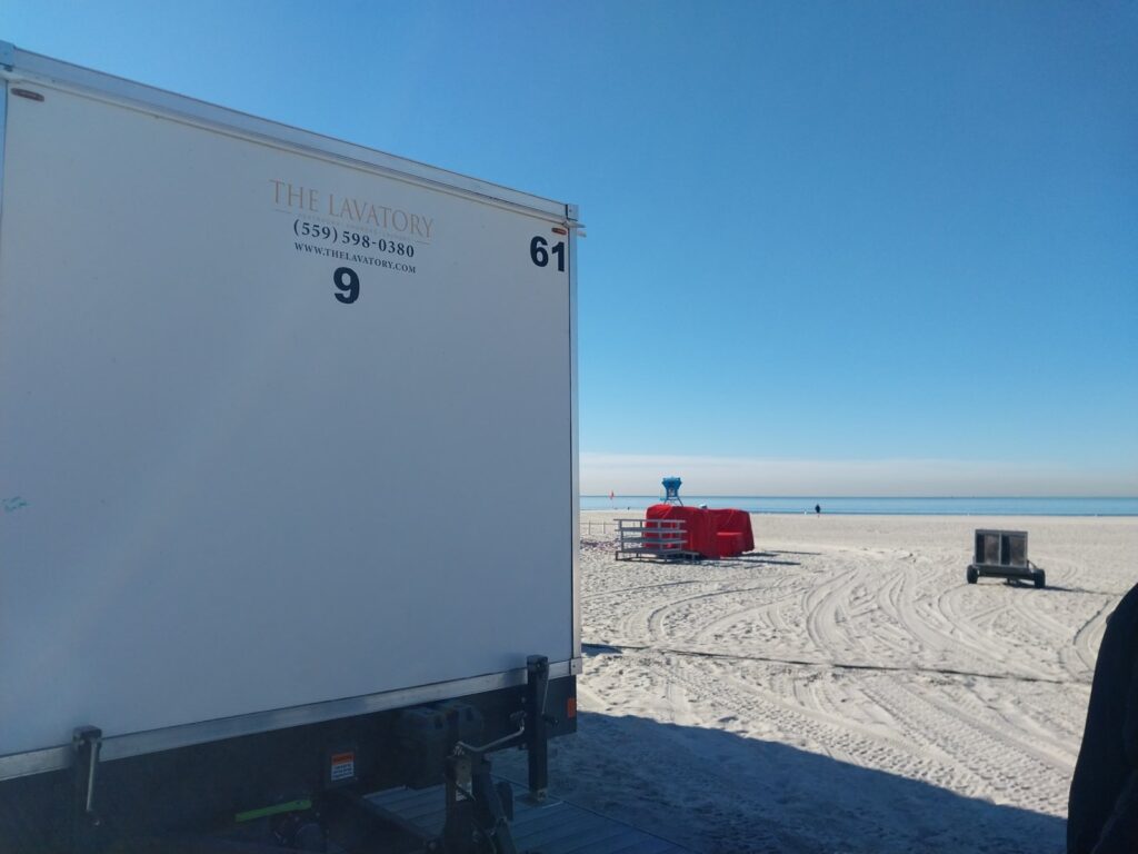 9-station bathroom trailer staged at the edge of a wide sandy beach with a lifeguard tower in the distance, The Lavatory Los Angeles