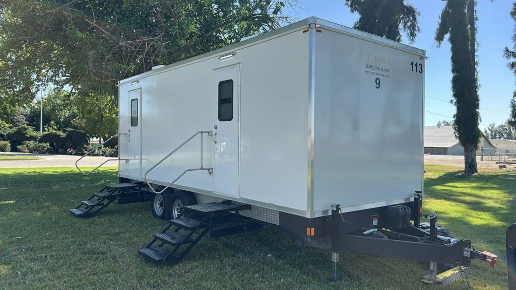 9-station bathroom trailer on a shaded park lawn with steps at each door, The Lavatory Los Angeles