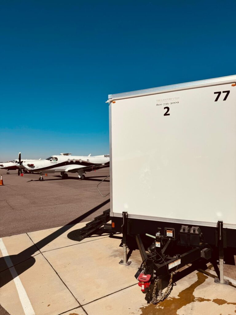 2-station bathroom trailer set on an airfield apron next to parked turboprop airplanes under a clear blue sky, The Lavatory Los Angeles