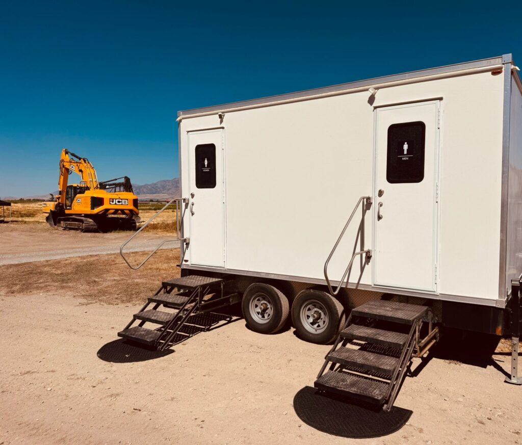2-station luxury bathroom trailer with women's and men's doors and steel stairs on a dirt job site, a yellow excavator and desert mountains behind it, The Lavatory Los Angeles