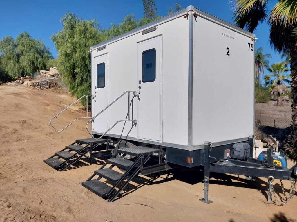 2-station bathroom trailer on leveling jacks on a sloped dirt site with a small generator on its tongue, The Lavatory Los Angeles