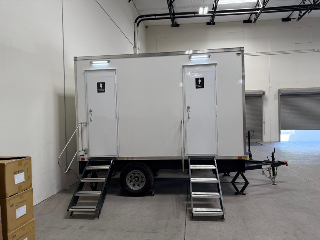 2-station bathroom trailer with lit door canopies parked inside a large warehouse beside roll-up dock doors, The Lavatory Los Angeles