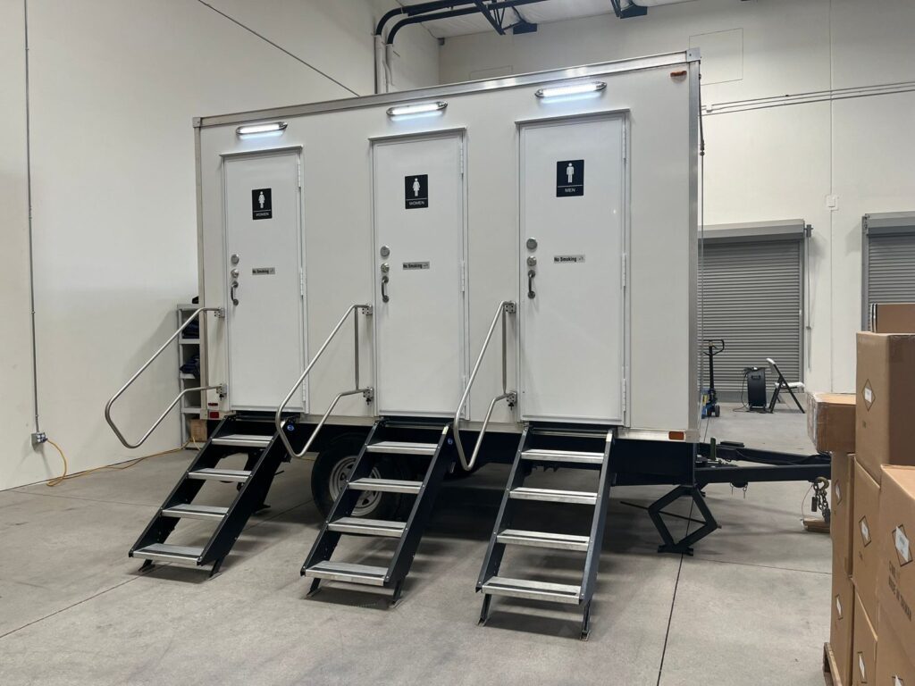 3-station bathroom trailer with three lit doors and stairs inside a distribution center beside stacked boxes, The Lavatory Los Angeles
