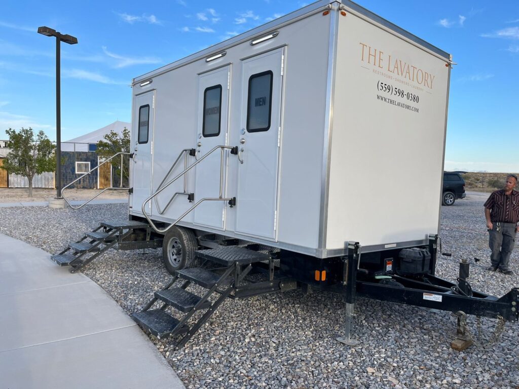 Site staff member standing beside a 3-station luxury bathroom trailer set on a gravel pad next to a walkway, The Lavatory Los Angeles