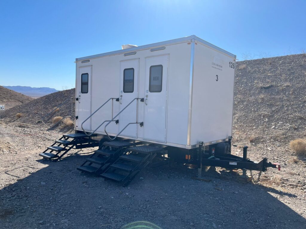 3-station bathroom trailer with three doors and handrails on a rocky desert lot below a gravel hillside, The Lavatory Los Angeles