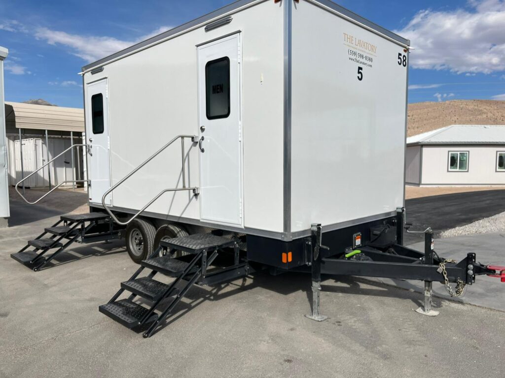 5-station bathroom trailer on blacktop at an industrial yard with desert hills and a metal carport behind it, The Lavatory Los Angeles