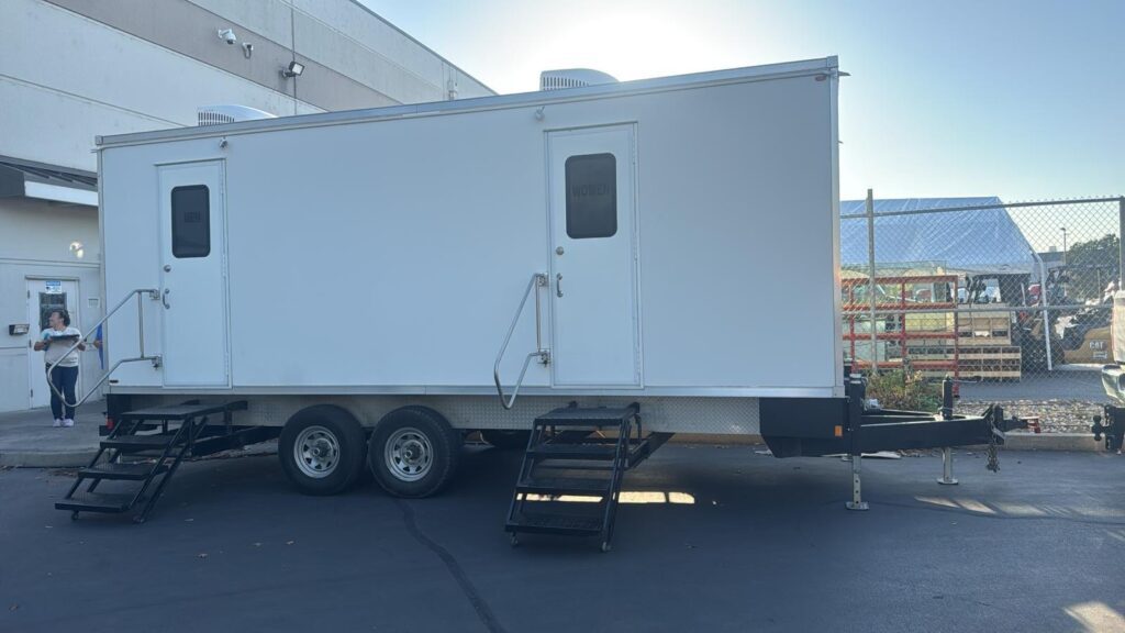 Worker walking out of a warehouse door beside a long bathroom trailer with two entry stairs on the parking lot, The Lavatory Los Angeles