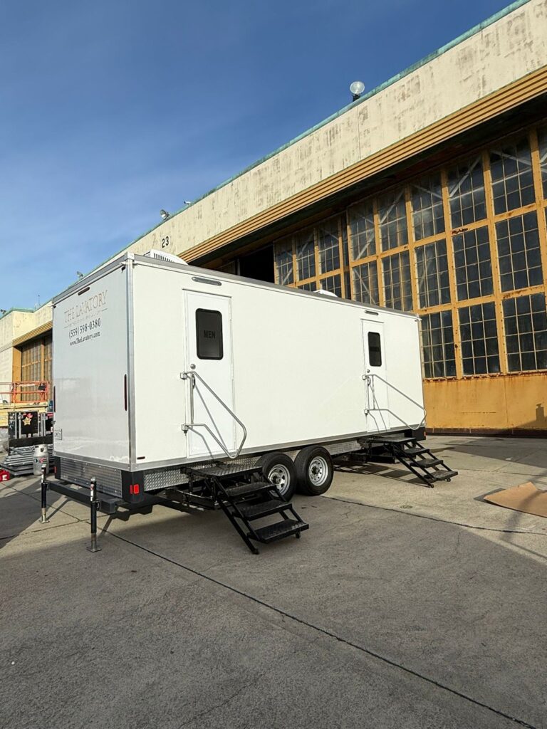 Long deluxe bathroom trailer parked on concrete beside the tall steel-framed windows of an old aircraft hangar, The Lavatory Los Angeles