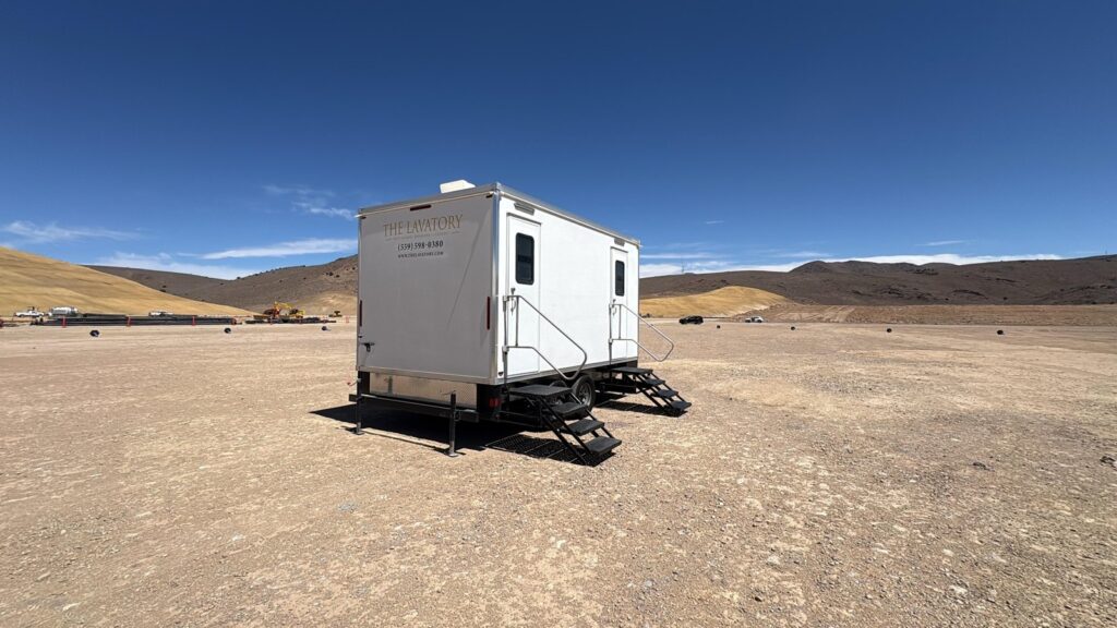 Portable bathroom trailer parked alone on a wide open desert dirt lot with brown hills on the horizon, The Lavatory Los Angeles