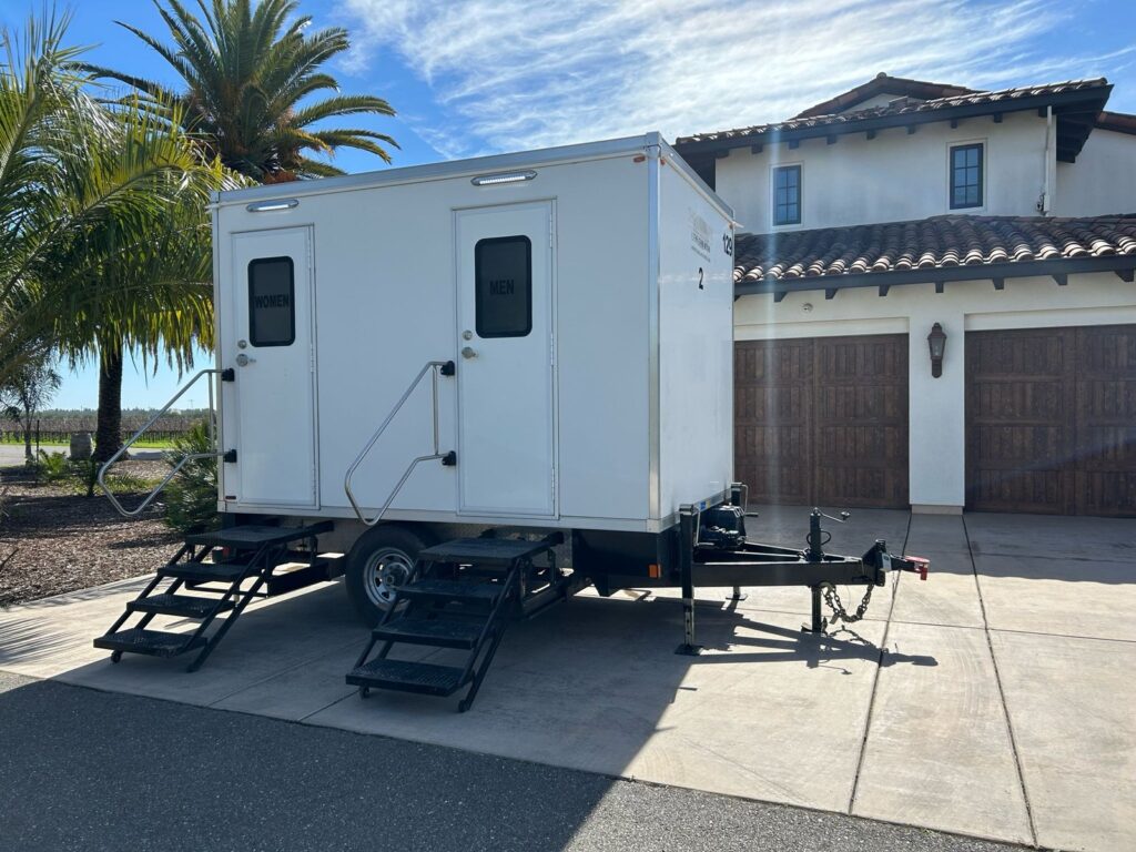 2-station bathroom trailer with women's and men's doors on the driveway of a Spanish style home with palm trees, The Lavatory Los Angeles