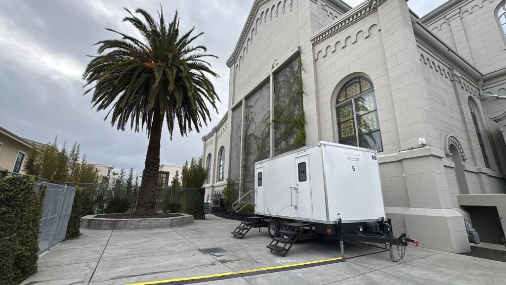5-station bathroom trailer set in a paved courtyard beside a stone church with stained glass windows and a palm tree, The Lavatory Los Angeles