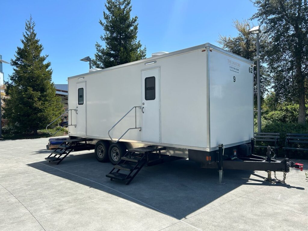 9-station bathroom trailer with two sets of steps on a paved campus plaza beside pine trees and benches, The Lavatory Los Angeles