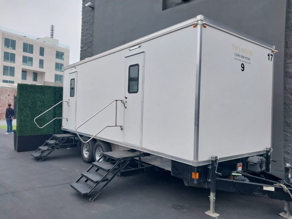 9-station bathroom trailer beside a hedge wall at an urban event venue as a guest walks by, The Lavatory Los Angeles