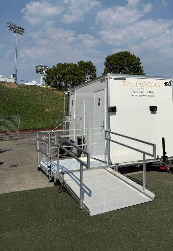 ADA bathroom trailer with a railed ramp on turf beside a stadium field and light tower, The Lavatory Los Angeles