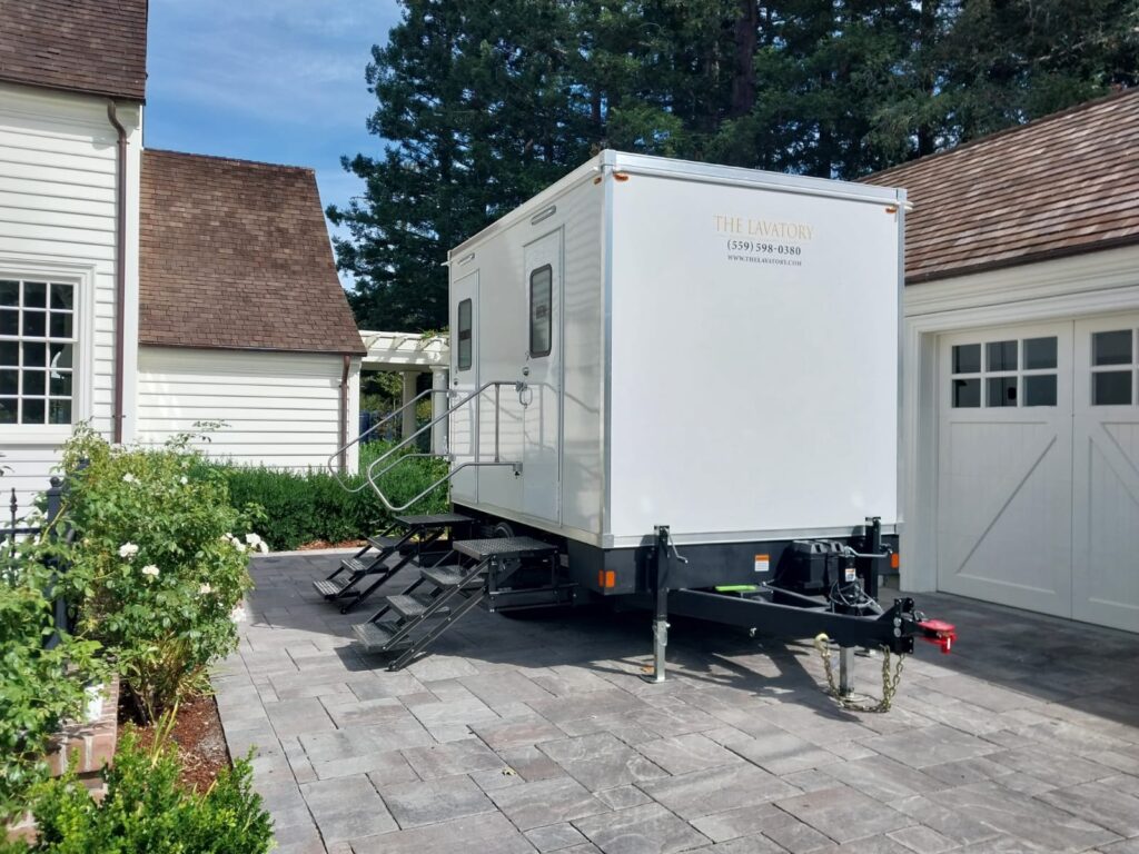 Bathroom trailer parked on a paver driveway between a white house and garage with rose bushes, The Lavatory Los Angeles