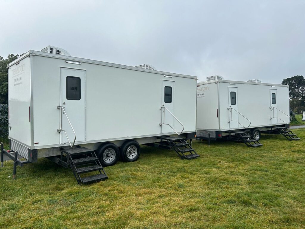 Two long bathroom trailers with men's and women's doors and steps set on a grass field, The Lavatory Los Angeles