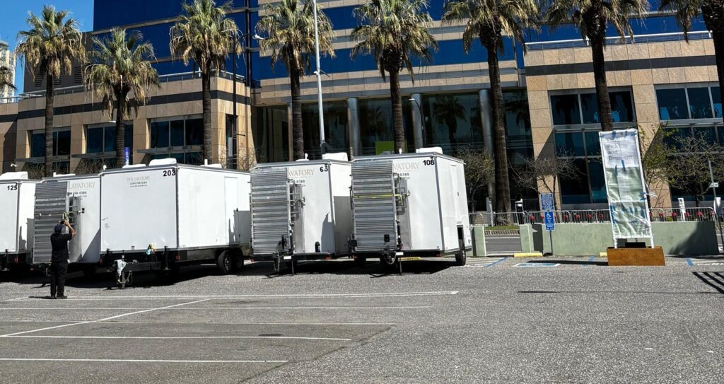 Worker setting up a long row of bathroom trailers in a downtown parking lot lined with palm trees, The Lavatory Los Angeles
