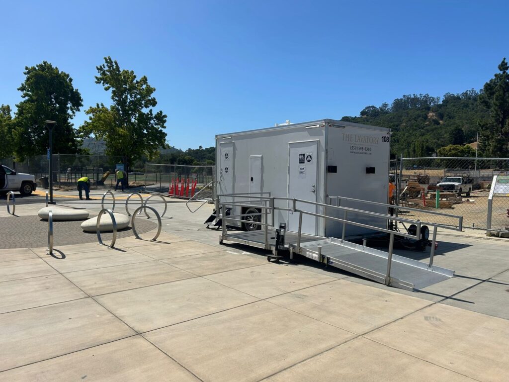 Workers in safety vests on a public plaza near an ADA plus 2 bathroom trailer with a railed ramp, The Lavatory Los Angeles
