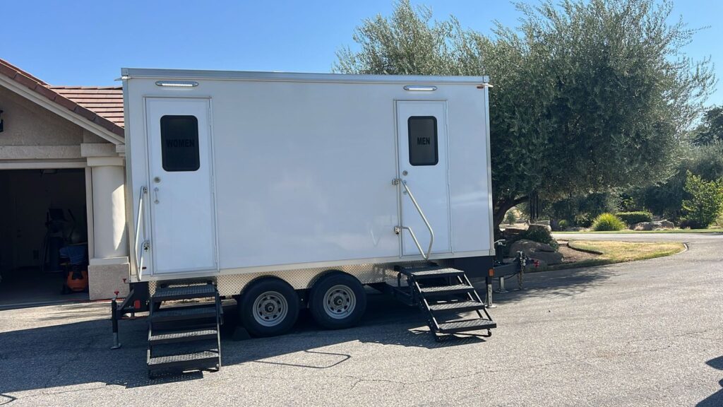 2-station bathroom trailer with women's and men's doors parked in a home driveway beside a tile roof garage and an olive tree, The Lavatory Los Angeles