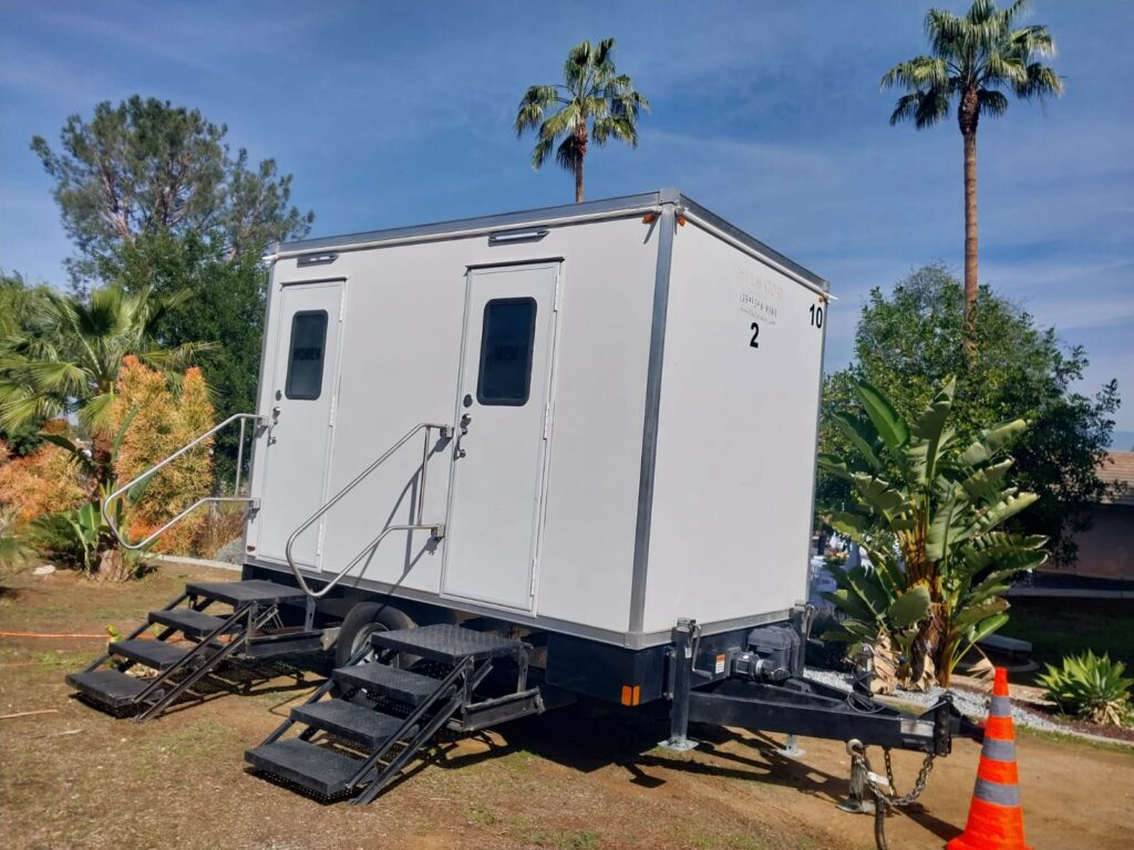 2-station bathroom trailer with steps at both doors set on dirt in a garden of palms and tropical plants beside a safety cone, The Lavatory Los Angeles