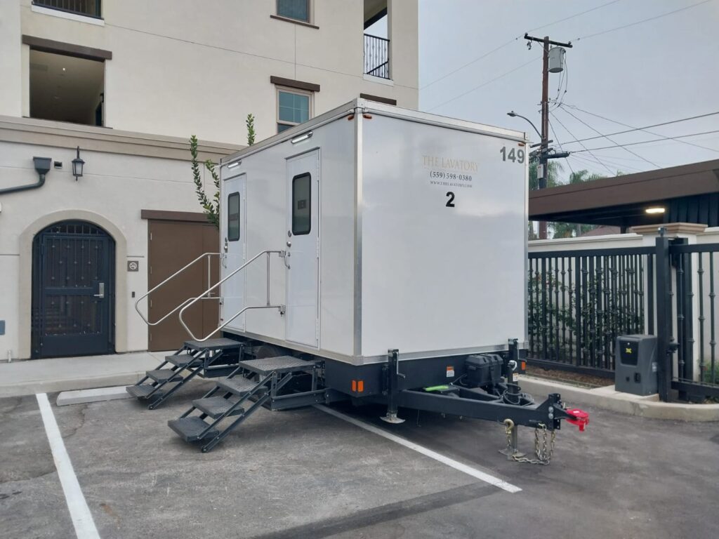 2-station bathroom trailer with steps at both doors parked beside a cream stucco building with an arched iron door, The Lavatory Los Angeles