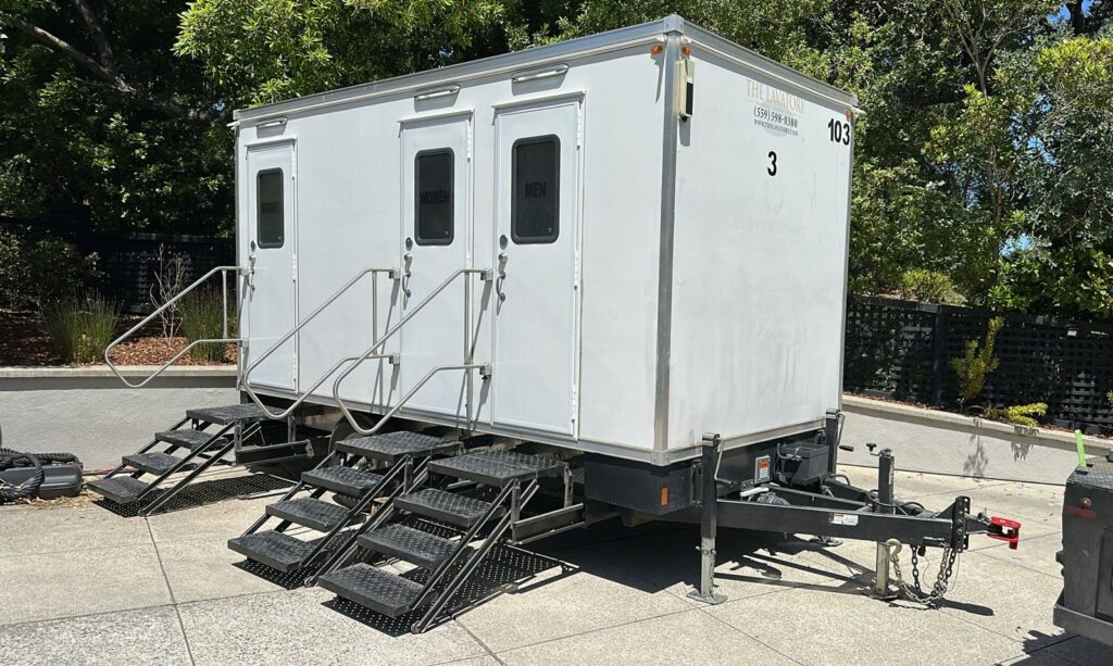 3-station bathroom trailer with three doors and steps set level on a concrete patio surrounded by trees, The Lavatory Los Angeles