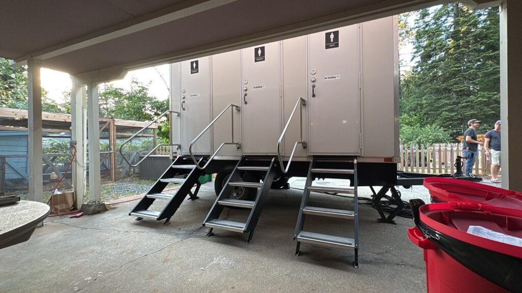 3-station portable bathroom trailer with three doors and steps set beside a covered patio while two people stand near the fence, The Lavatory Los Angeles