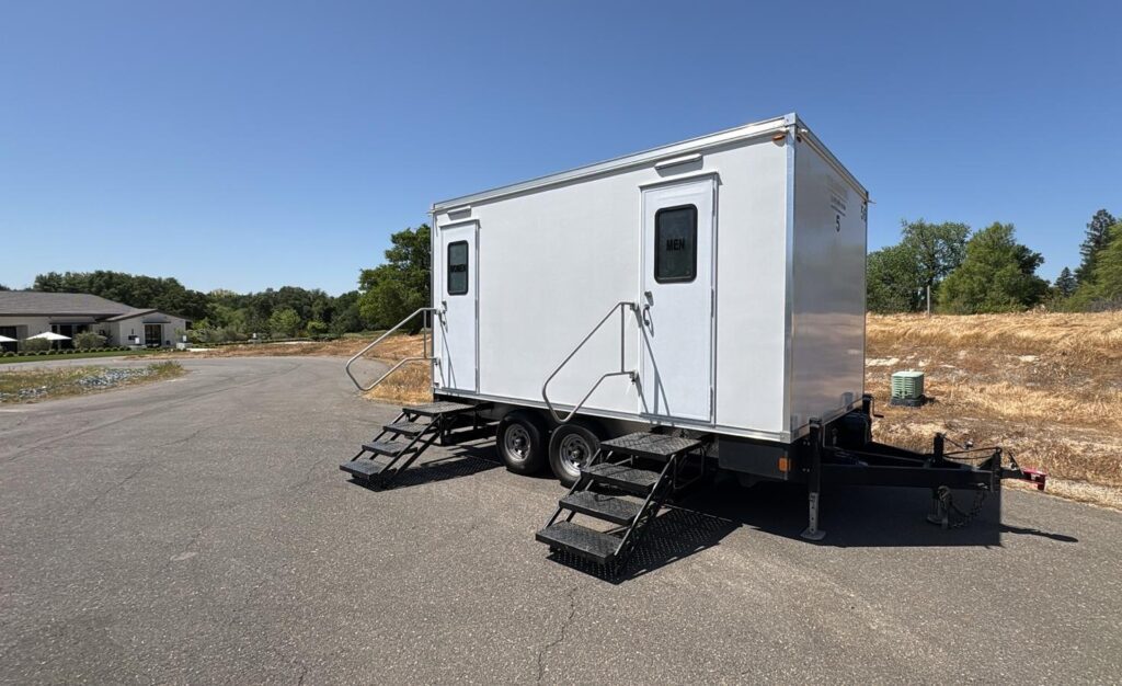5-station bathroom trailer parked on a paved street at a new subdivision site with dry grass lots behind it, The Lavatory Los Angeles