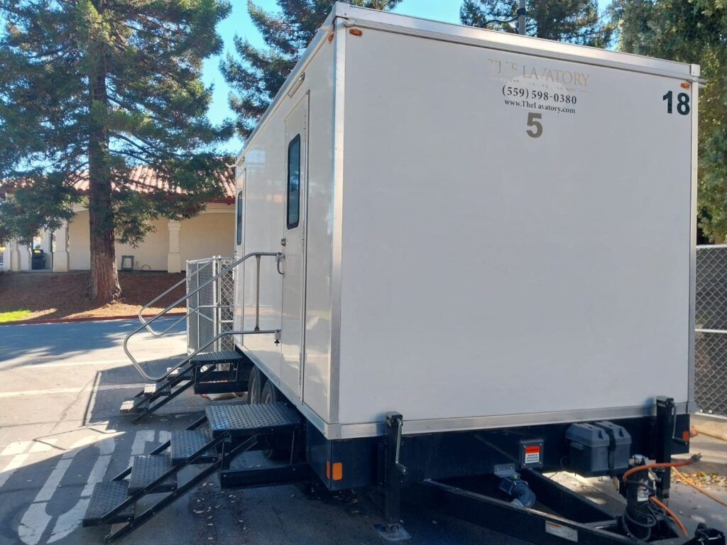 5-station bathroom trailer with steps and handrails parked in a lot beside a tile roof building and tall pine trees, The Lavatory Los Angeles