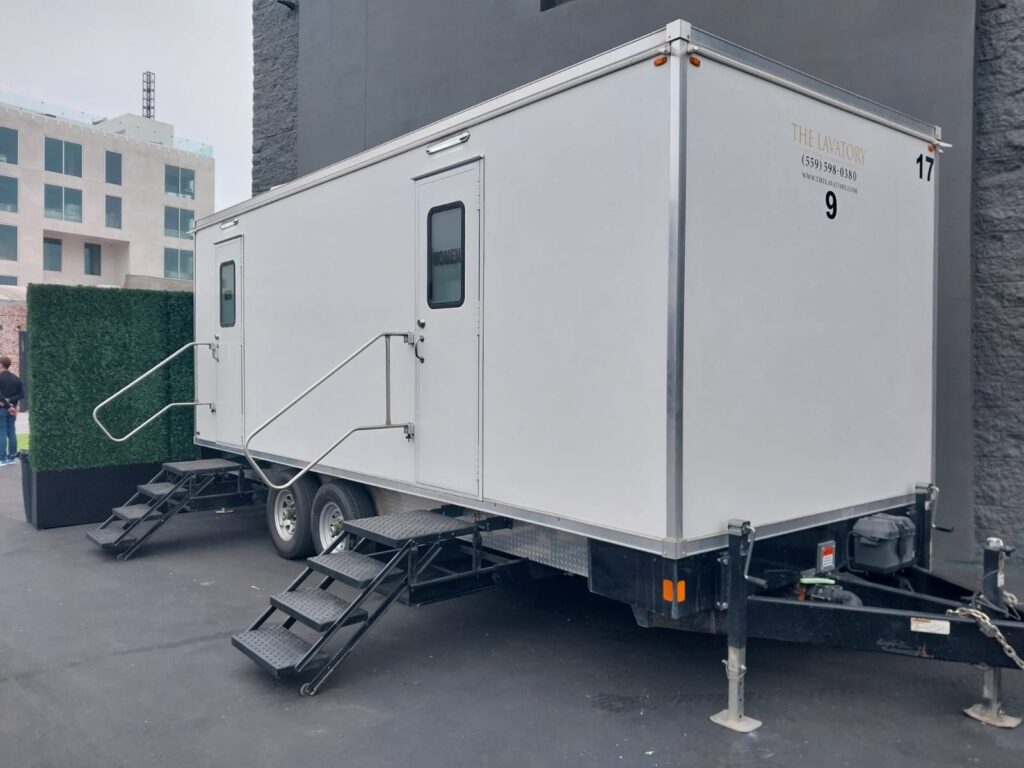 9-station luxury bathroom trailer with two entry doors and fold-down steps parked beside a gray building and a green hedge wall, with a guest standing nearby, The Lavatory Los Angeles
