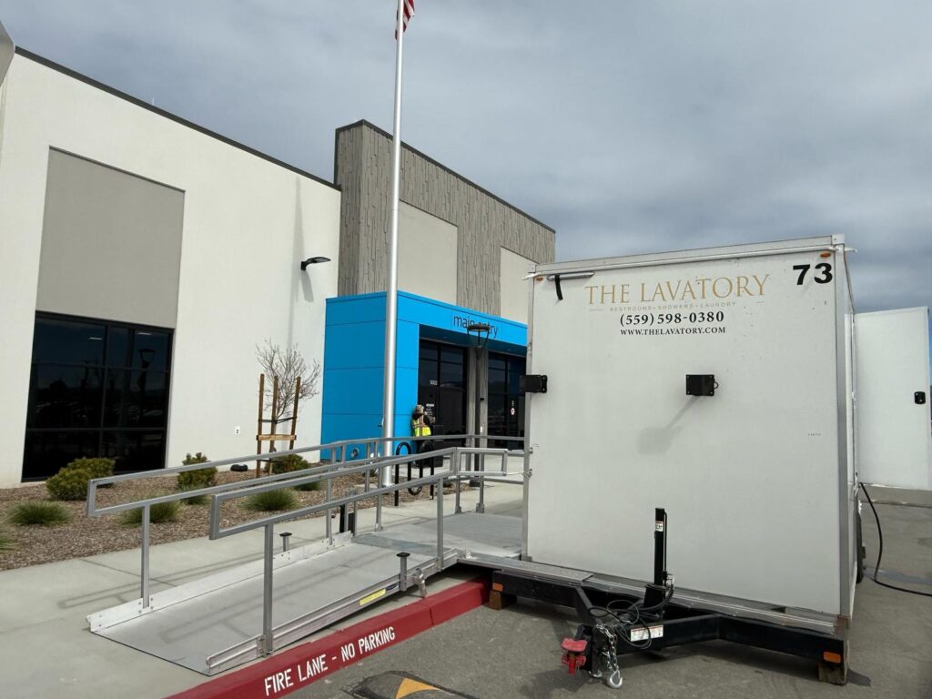 ADA plus 2 bathroom trailer with an aluminum wheelchair ramp at an office building entrance, with a worker in a safety vest at the door, The Lavatory Los Angeles