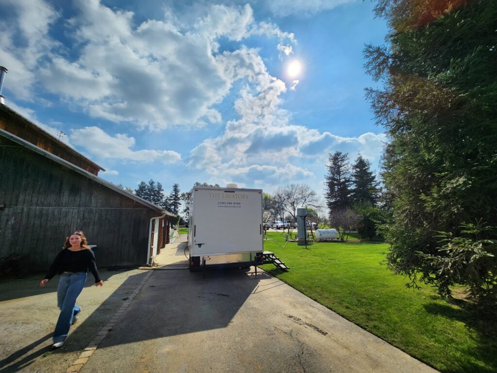 A guest walks up a driveway past a wooden barn, with a bathroom trailer parked at the edge of a green lawn behind her, The Lavatory Los Angeles