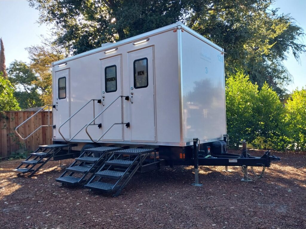 3-station bathroom trailer with three sets of railed steps set on mulch in a shaded backyard, The Lavatory Los Angeles
