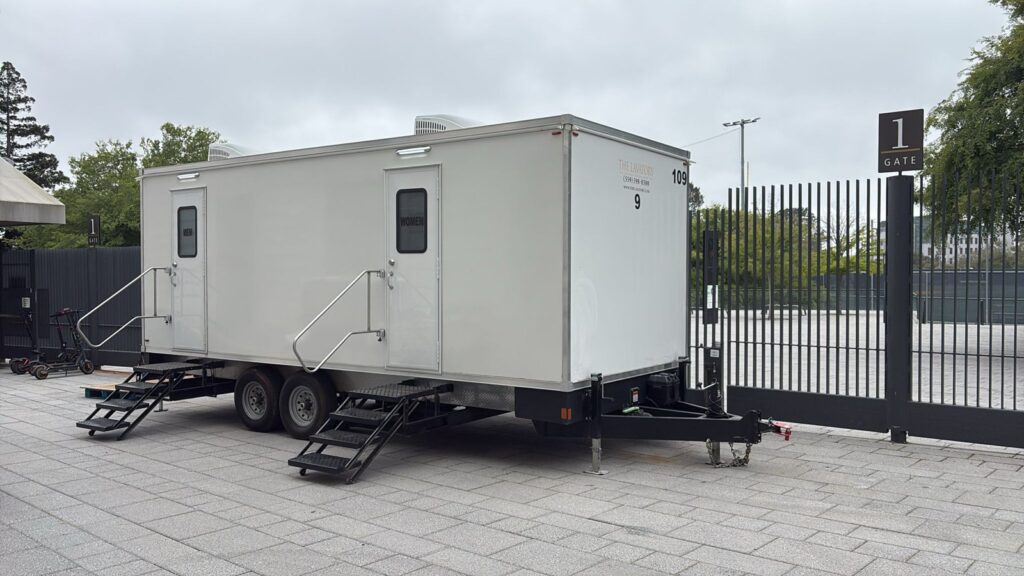 9-station bathroom trailer on a paver courtyard beside a numbered entry gate at an event venue, The Lavatory Los Angeles