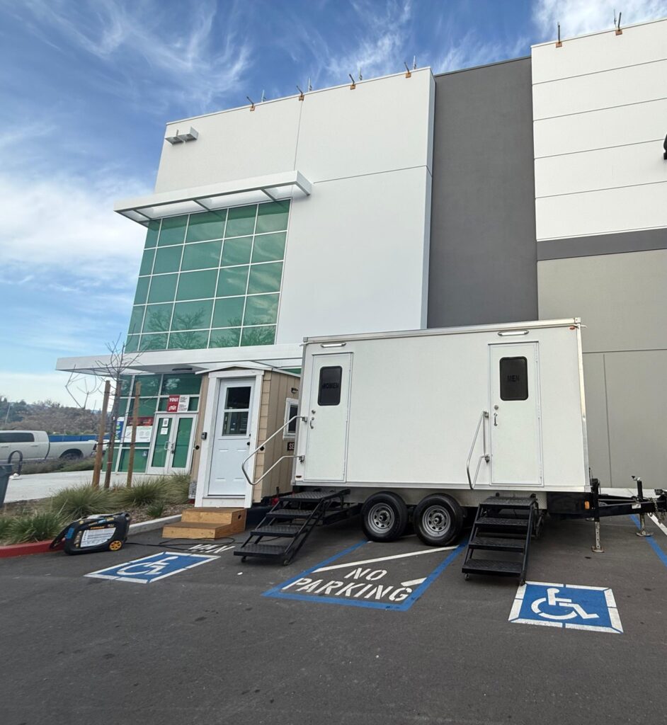 Bathroom trailer with two doors and steps beside the office entrance of a new warehouse building, The Lavatory Los Angeles