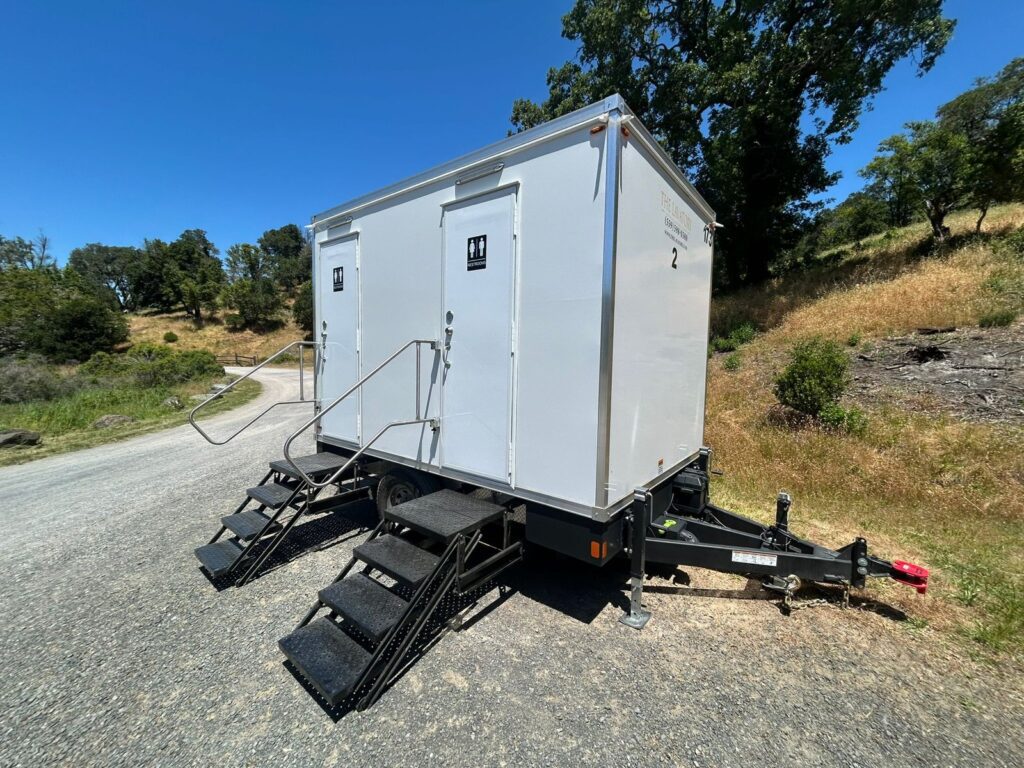 2-station bathroom trailer with fold-down steps parked on a gravel road beside oak trees and dry grass hills, The Lavatory Los Angeles