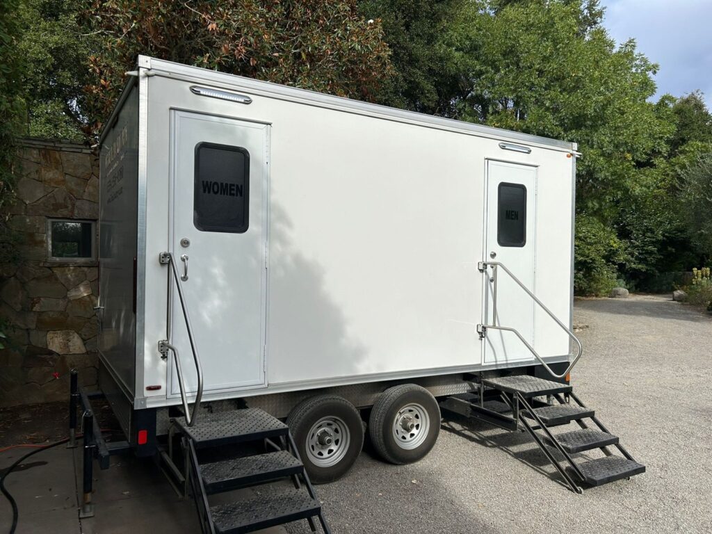 Bathroom trailer with women's and men's doors parked on a paved lot beside a stone wall and trees, The Lavatory Los Angeles