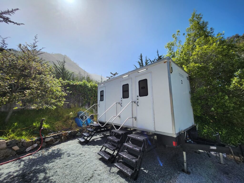 3-station bathroom trailer with three sets of steps on a gravel pad below a green hillside, The Lavatory Los Angeles