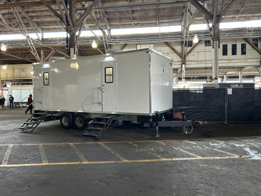 9-station bathroom trailer parked inside a large stage building as production staff walk past, The Lavatory Los Angeles