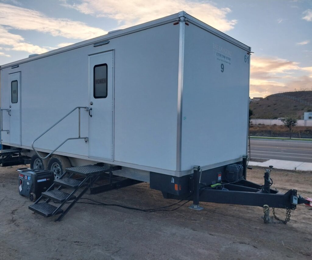 9-station bathroom trailer with a generator on a dirt lot beside a new road and a graded hillside at sunset, The Lavatory Los Angeles