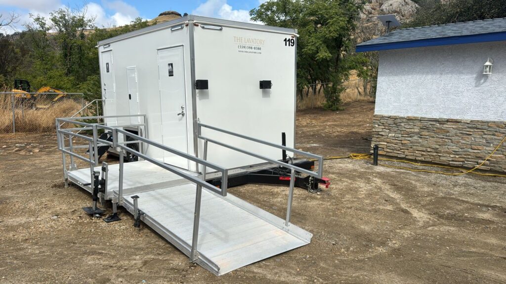ADA plus 2 bathroom trailer with ramp on a dirt site with an excavator and rocky hills behind it, The Lavatory Los Angeles