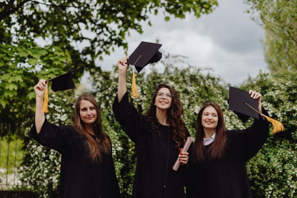 Three graduates in black gowns raising their caps outdoors under the trees, The Lavatory Los Angeles