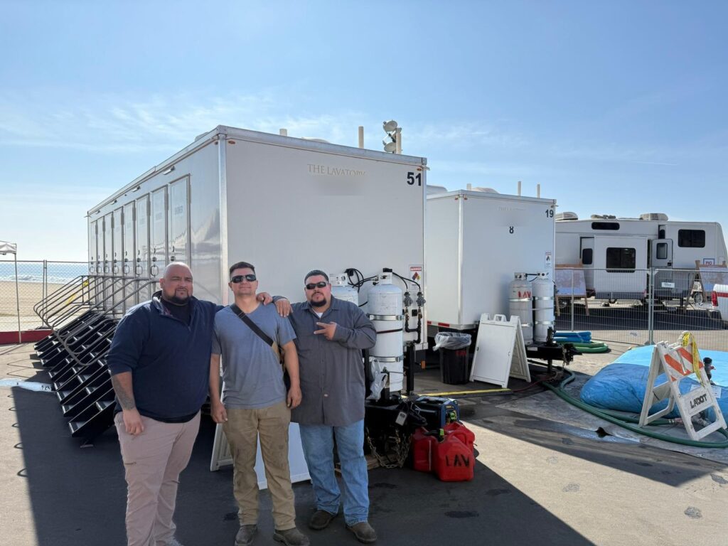 Three members of our service team standing in front of shower trailers they set up on a beach parking lot, The Lavatory Los Angeles