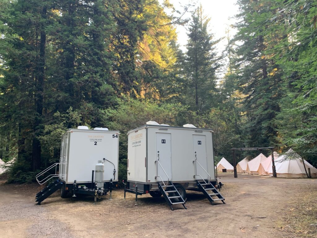 Shower and bathroom trailers with steps set up in the forest beside rows of bell tents at a camp, The Lavatory Los Angeles