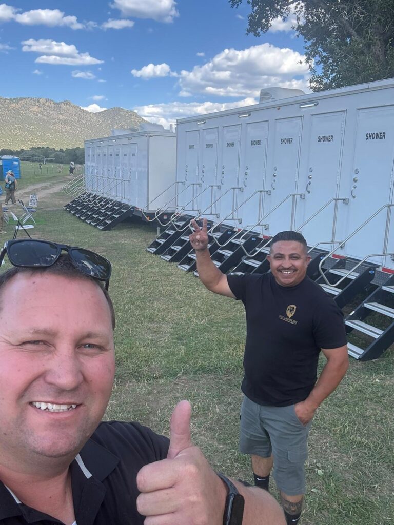 Two members of our team smiling in front of a row of 8-station shower trailers at a large event, The Lavatory Los Angeles