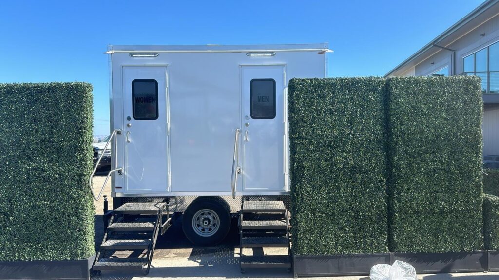 2-station bathroom trailer framed by green hedge panels at an outdoor event, The Lavatory Los Angeles
