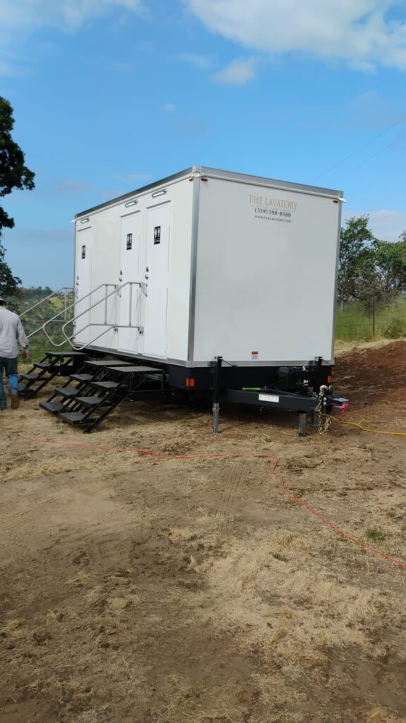 3-station bathroom trailer leveled on a dirt pad with a worker standing beside the steps, The Lavatory Los Angeles