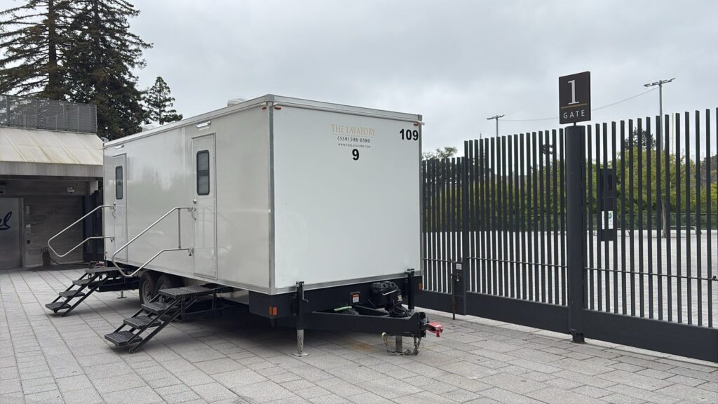 9-station bathroom trailer on a paver plaza beside a tall steel gate, The Lavatory Los Angeles